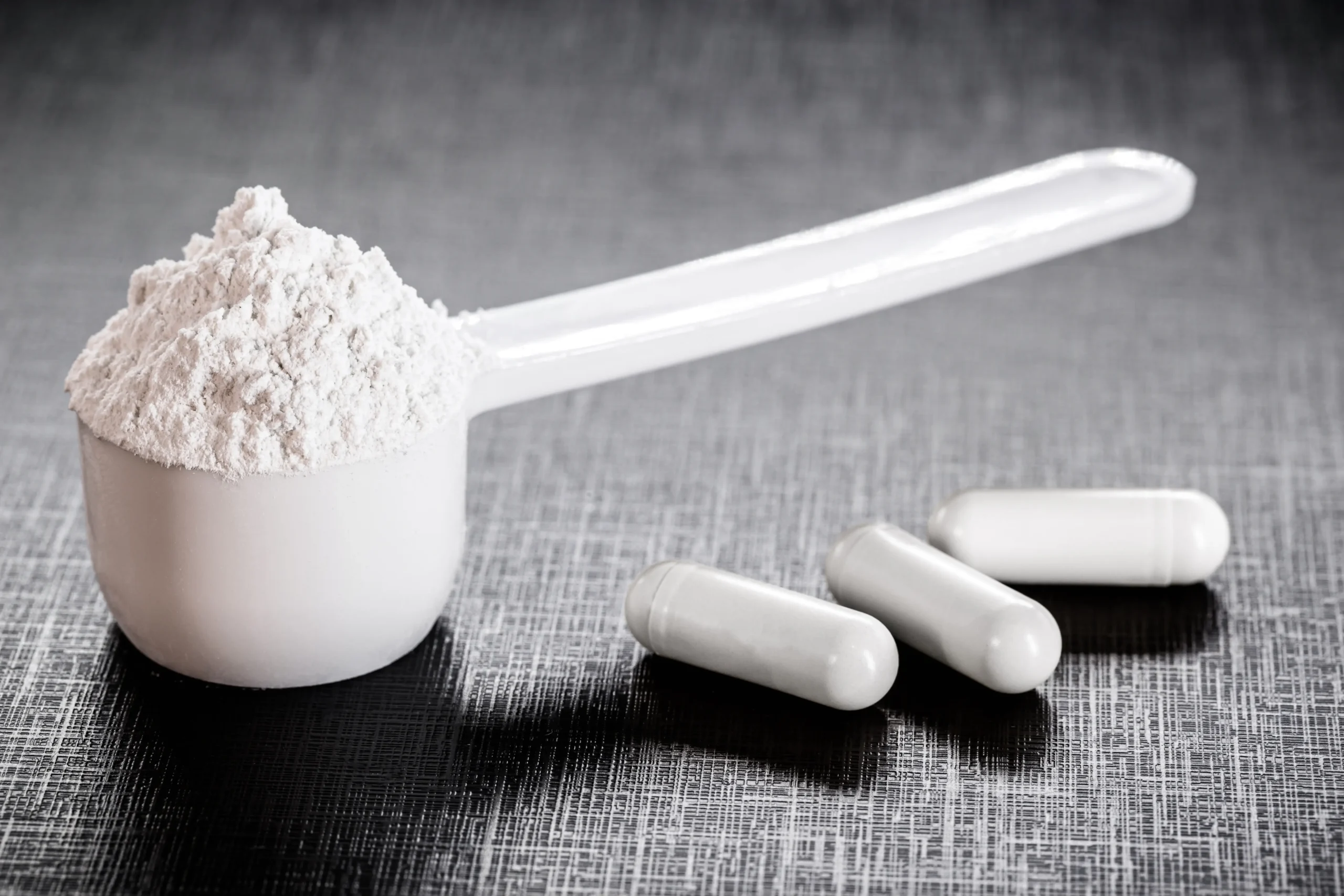 A close-up image of a scoop filled with creatine placed on a textured gray surface, with three white capsules lying next to it.