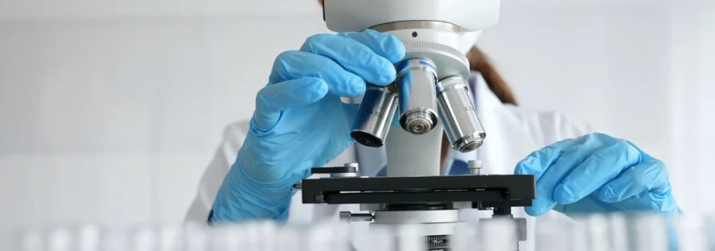 Laboratory scientist examining a sample through a microscope while wearing blue protective gloves.