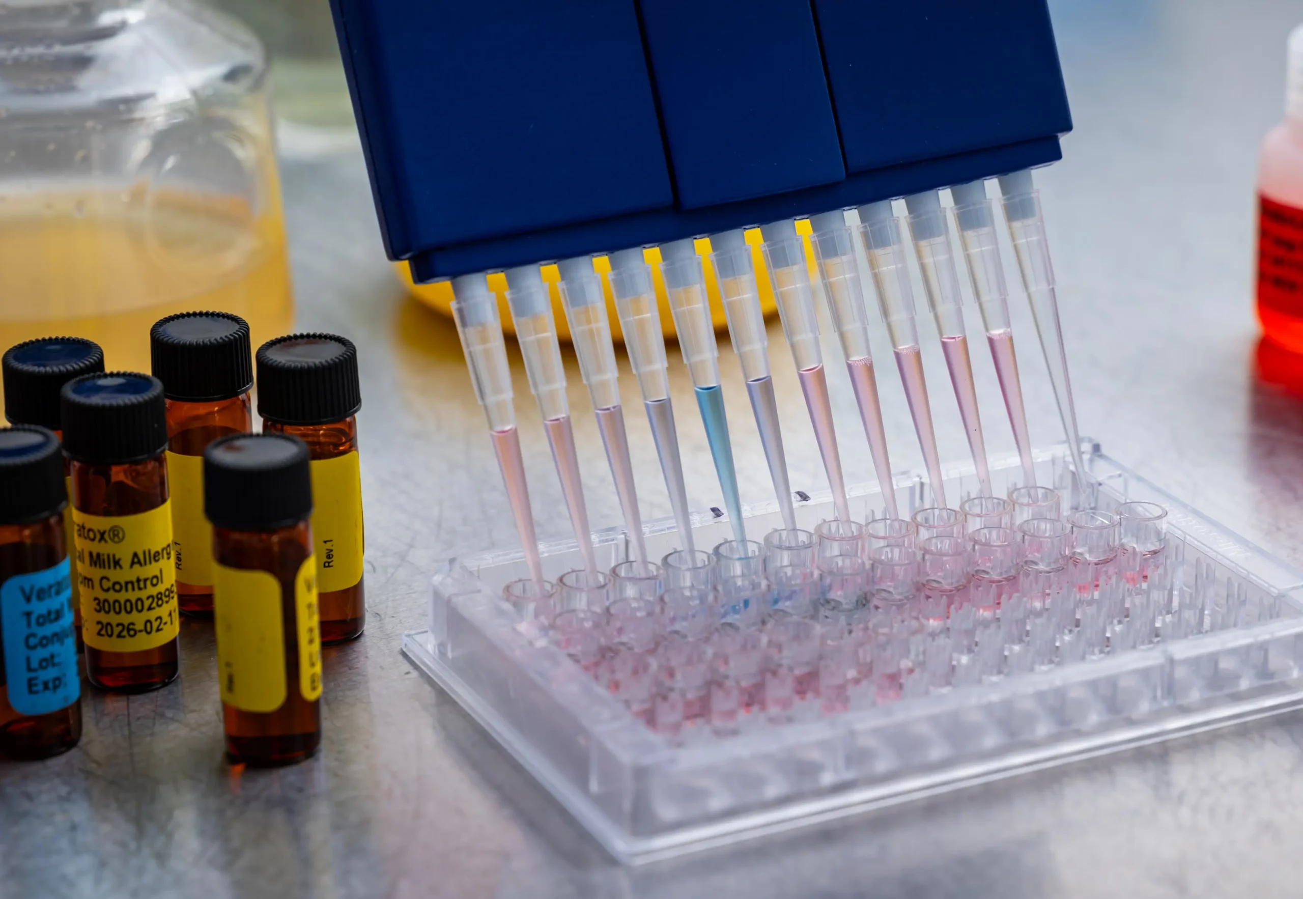 Laboratory pipette system transferring liquid samples into a multi-well plate beside labeled laboratory sample bottles.